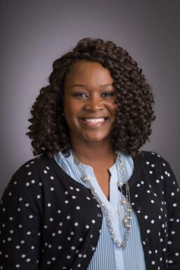 Staff photograph in Tampa, Fl of smiling, curly haired woman with polka dotted black sweater against gray.